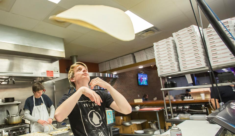 Kitchen scene with pizza tossed in the air by young man in black t-shirt.