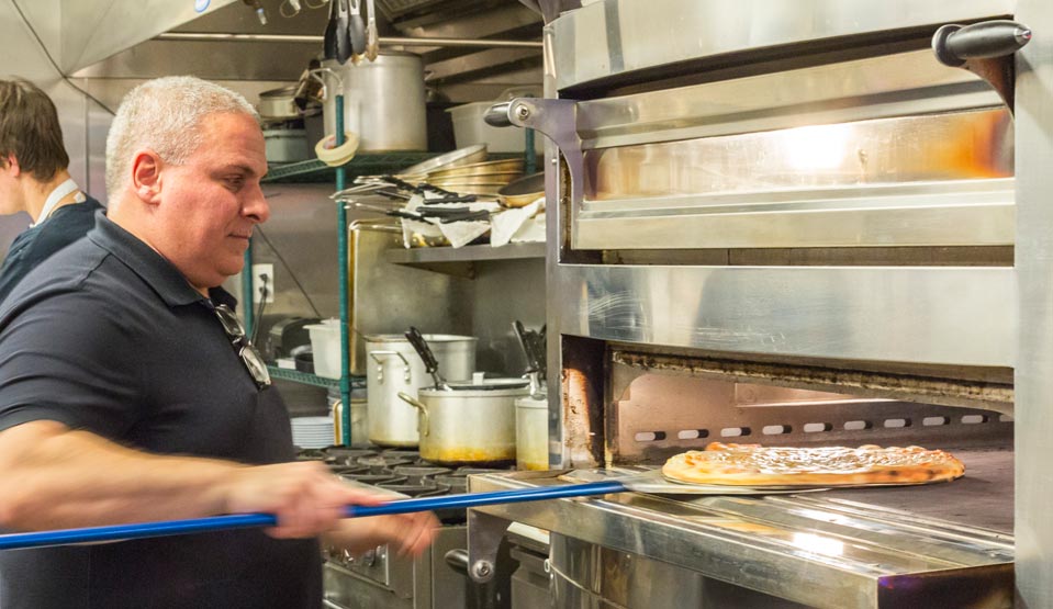 Ed Bisconti, owner of Borgata Pizza Cafe sliding a pizza on a peel into a large metal pizza oven.