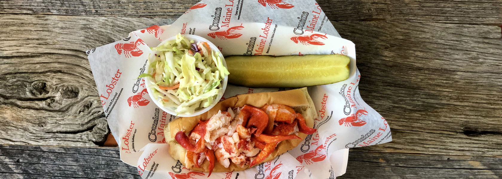 Close-up view of Cousins Maine Lobster Connecticut Lobster Roll with cup of coleslaw and pickle slice in basket.