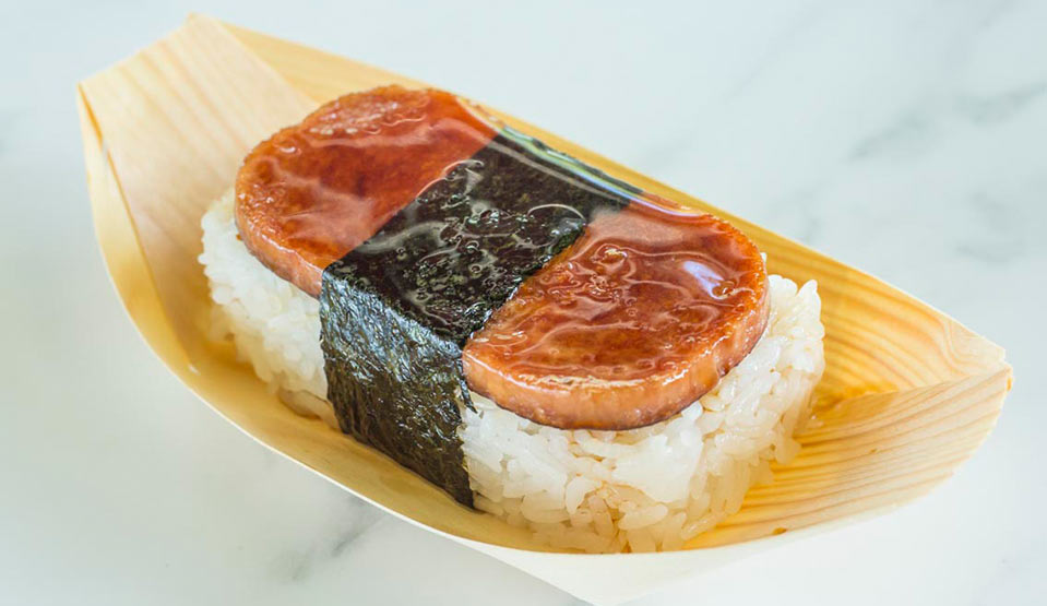 Slice of raw fish on top of rectangular rice bed wrapped with strip of seaweed in bamboo paper dish.