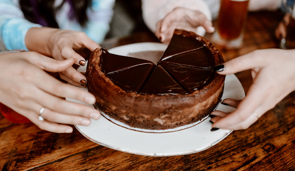 Chocolate-covered cheesecake on a plate with hands selecting a piece.