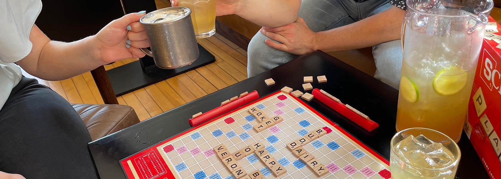 Hands toasting with cocktails above Scrabble game board in play.